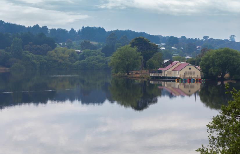Daylesford lake, Victoria. Peaceful lakeside scene with a boathouse reflected in still water, surrounded by lush green trees and hills under a cloudy sky, conveying tranquility.