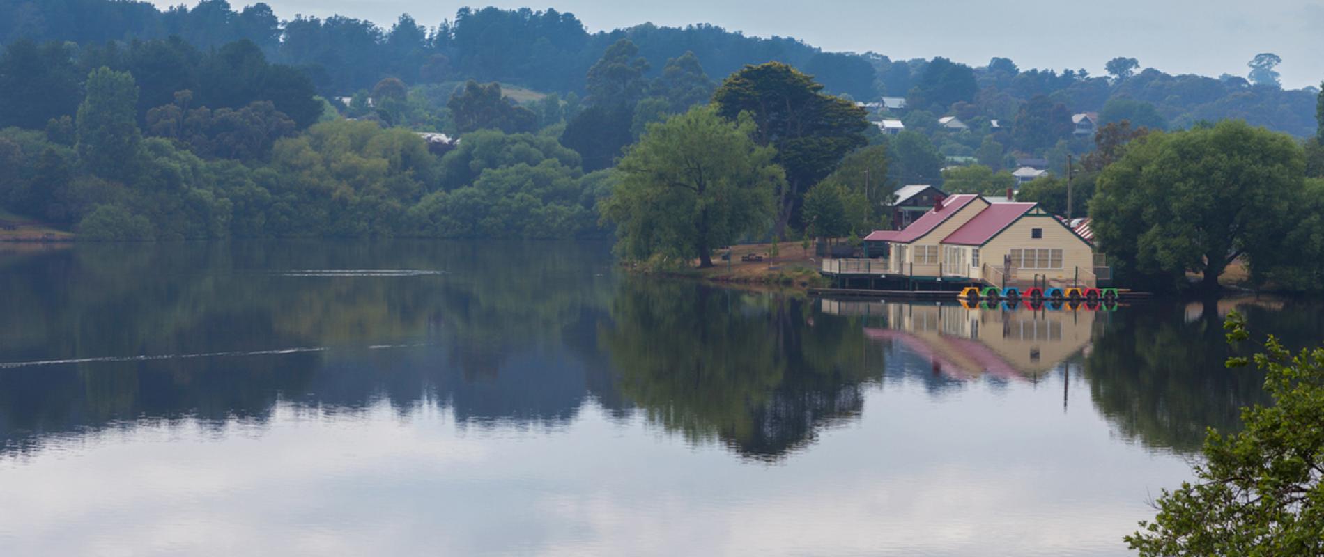 Daylesford lake, Victoria. Peaceful lakeside scene with a boathouse reflected in still water, surrounded by lush green trees and hills under a cloudy sky, conveying tranquility.